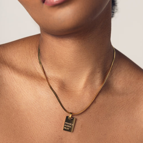 Close-up of a person wearing an 18k gold  plated herringbone necklace with a rectangular pendant that has engraved text reading "Phahla Pusha Pray