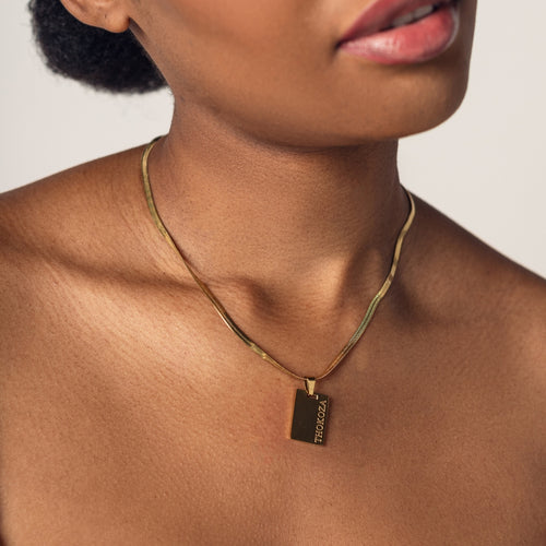 Close-up of a person wearing an 18K gold plated herringbone necklace with a rectangular pendant that has engraved text reading "Thokoza" 