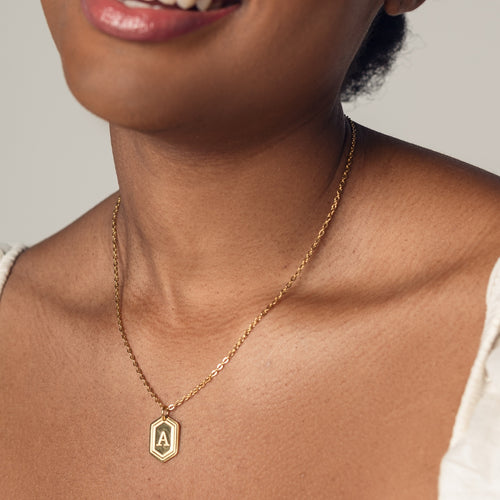 A close-up image of a woman wearing a delicate 18k gold plated chain necklace with a hexagonal pendant. The pendant features a bold, engraved letter "A" in the center. 