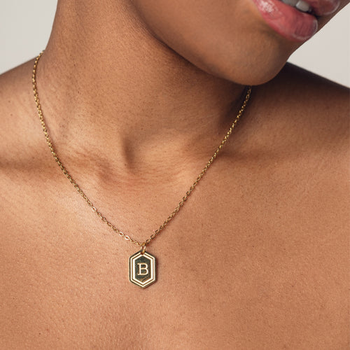 A close-up image of a woman wearing a delicate 18k gold plated chain necklace with a hexagonal pendant. The pendant features a bold, engraved letter "B" in the center. 