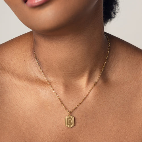 A close-up image of a woman wearing a delicate 18k gold plated chain necklace with a hexagonal pendant. The pendant features a bold, engraved letter "D" in the center. 