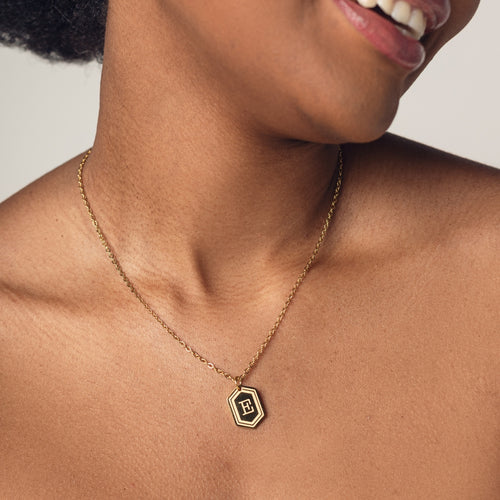 A close-up image of a woman wearing a delicate 18k gold plated chain necklace with a hexagonal pendant. The pendant features a bold, engraved letter "E" in the center. 