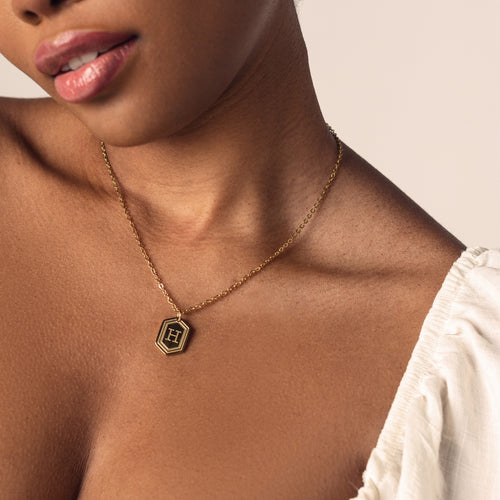 A close-up image of a person wearing a delicate 18k gold  plated chain necklace with a hexagonal pendant. The pendant features a bold, engraved letter "H" in the center. 