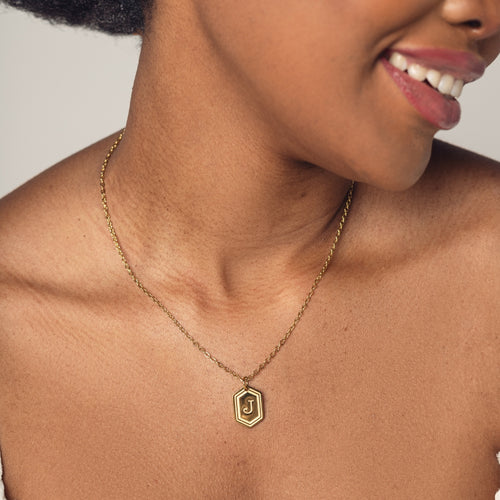 A close-up image of a woman wearing a delicate 18k gold plated chain necklace with a hexagonal pendant. The pendant features a bold, engraved letter "J" in the center. 