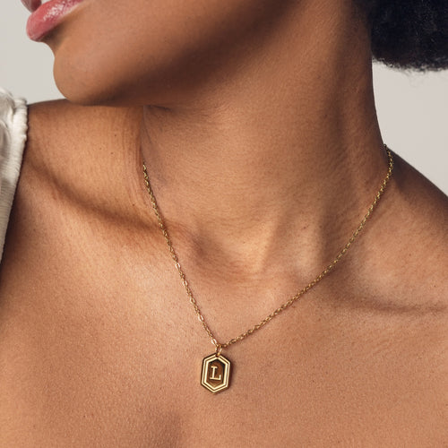 A close-up image of a woman wearing a delicate 18k gold plated chain necklace with a hexagonal pendant. The pendant features a bold, engraved letter "L" in the center. 