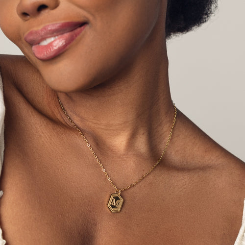 A close-up image of a woman wearing a delicate 18k gold  plated chain necklace with a hexagonal pendant. The pendant features a bold, engraved letter "M" in the center. 