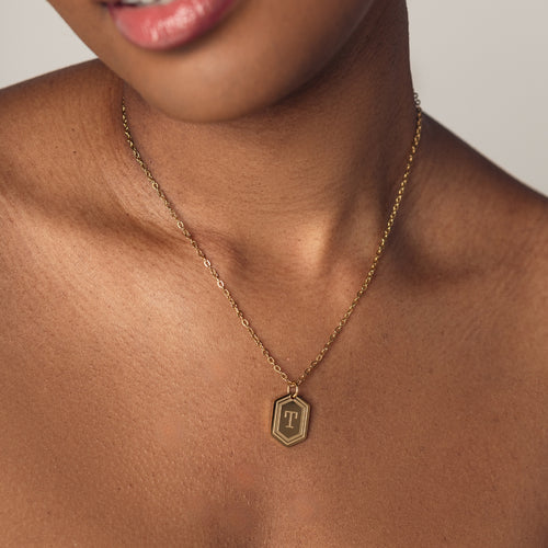 A close-up image of a woman wearing a delicate 18k gold plated chain necklace with a hexagonal pendant. The pendant features a bold, engraved letter "T" in the center. 