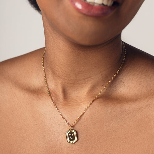 A close-up image of a woman wearing a delicate 18k gold plated chain necklace with a hexagonal pendant. The pendant features a bold, engraved letter "U" in the center. 