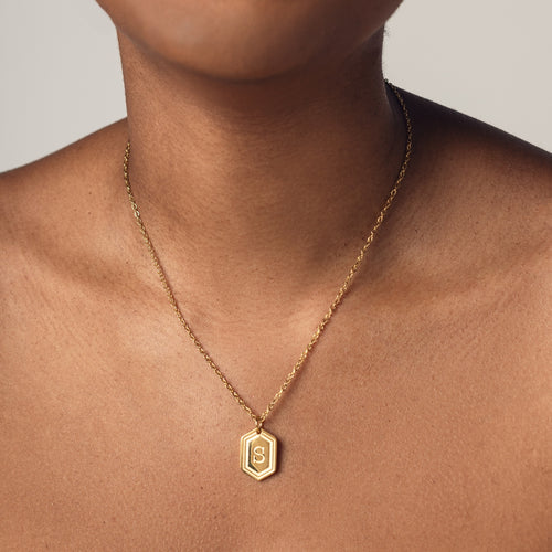 A close-up image of a person wearing a delicate 18k gold  plated chain necklace with a hexagonal pendant. The pendant features a bold, engraved letter "S" in the center. 